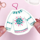 Decorative hand embroidered christmas jumper ornament with snowflake design held by a hand on a pink background