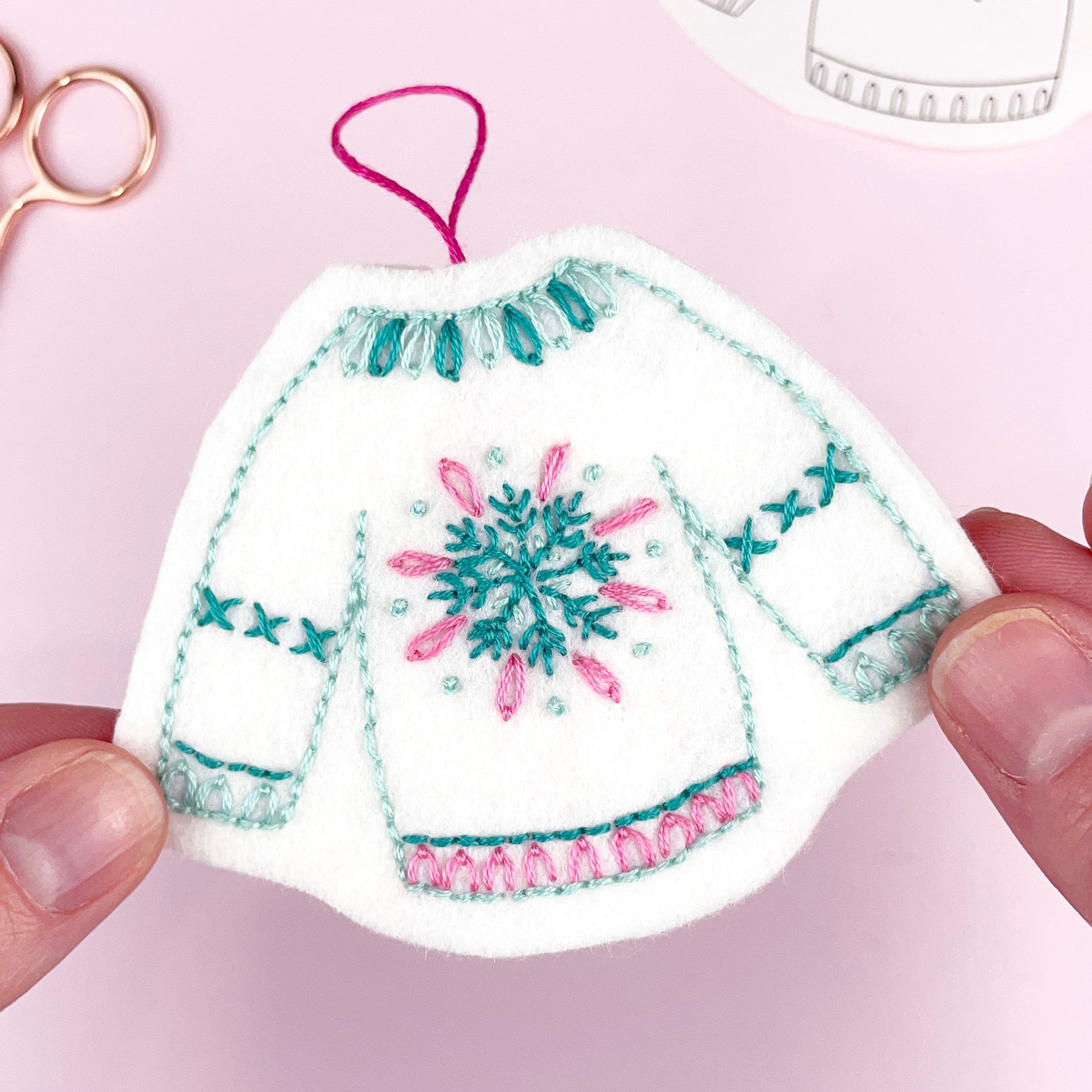 Decorative hand embroidered christmas jumper ornament with snowflake design held by a hand on a pink background