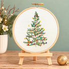 A finished embroidery piece showing a decorated Christmas tree inside a wooden hoop on a small wooden stand, set on a tabletop with a blurred holiday scene in the background.