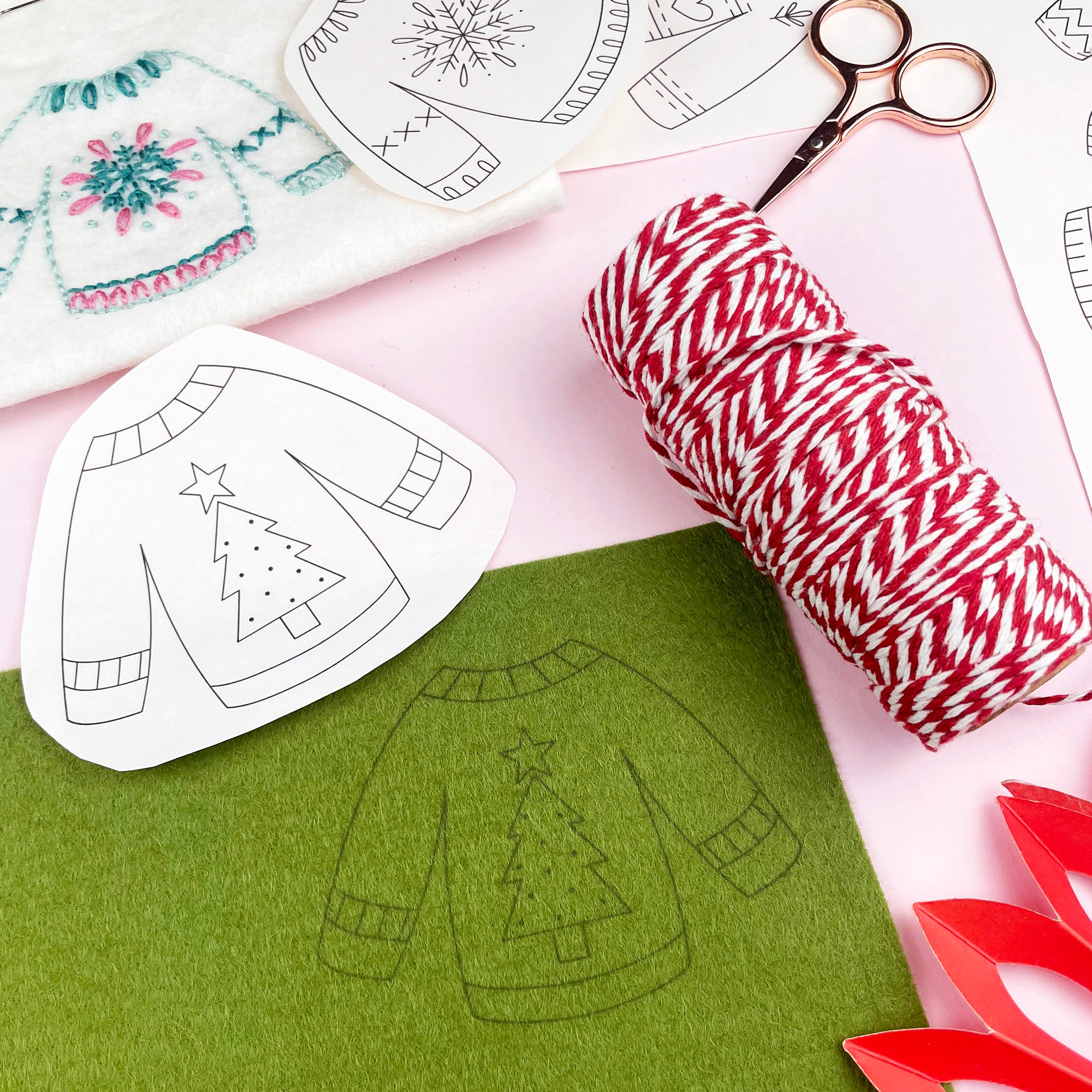 embroidery materials including paper patterns of Christmas sweaters, scissors, and red and white string on a pink surface.