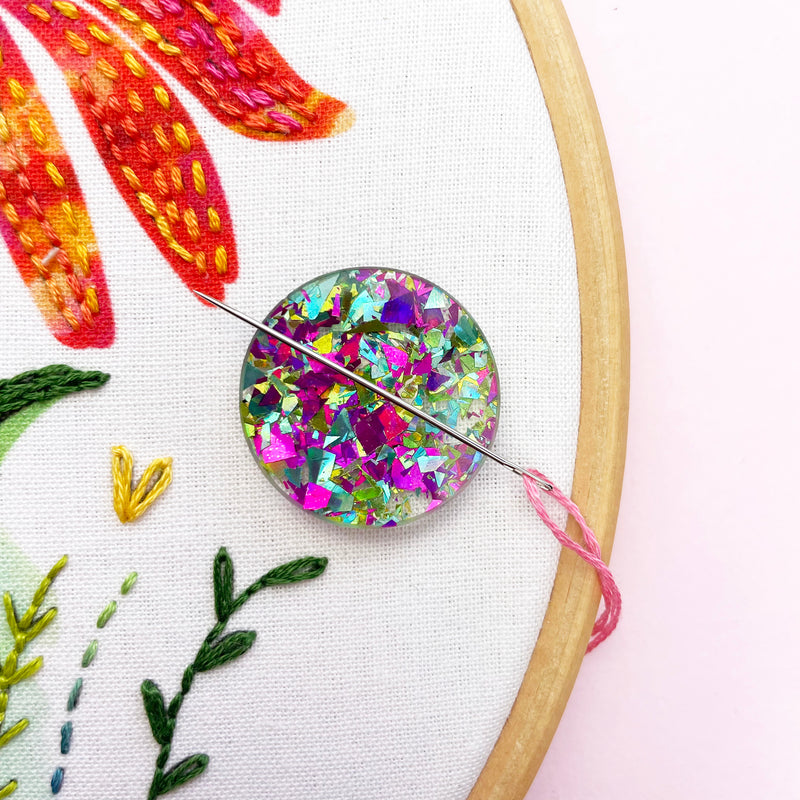 Multicolour glitter circle magnetic needle minder attached to embroidery fabric in wooden hoop with floral stitch design.