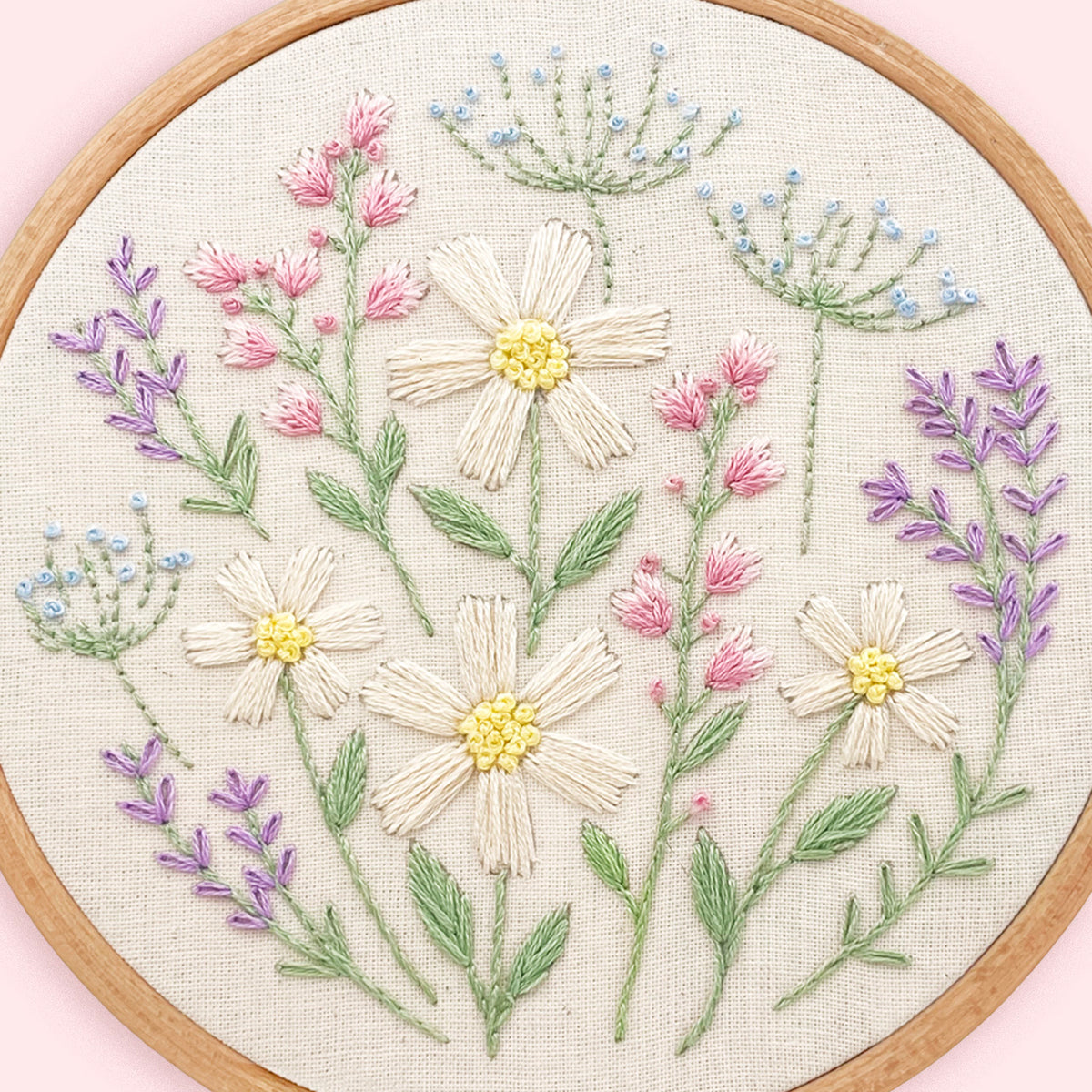 A circular embroidery hoop showcasing a detailed floral embroidery design on textured cream fabric. The design features cream-colored daisies with yellow centers, pink flowers, green stems and leaves, and delicate blue and lavender details.