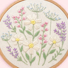 A circular embroidery hoop showcasing a detailed floral embroidery design on textured cream fabric. The design features cream-colored daisies with yellow centers, pink flowers, green stems and leaves, and delicate blue and lavender details.