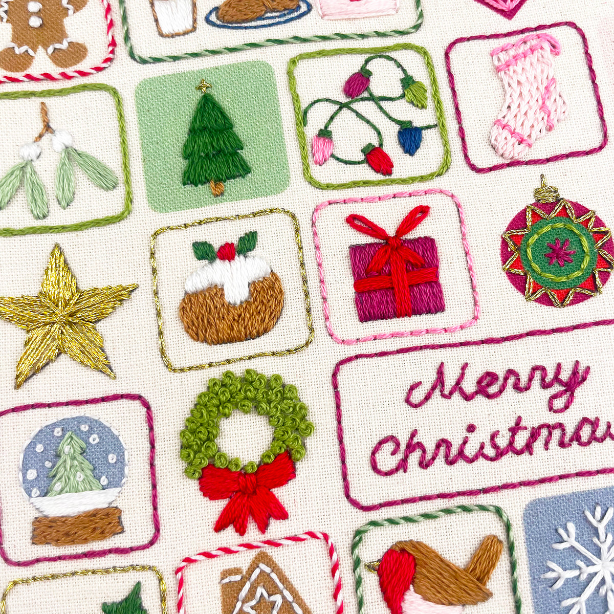 Close-up of a Christmas-themed embroidered advent calendar  featuring stitched motifs like a tree, stocking, lights, pudding, gift box, ornament, wreath, snow globe, and the words “Merry Christmas” on a cream woven background.