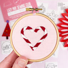 Embroidery hoop with candy cane heart design held by a hand, with a pink background and sewing club materials.