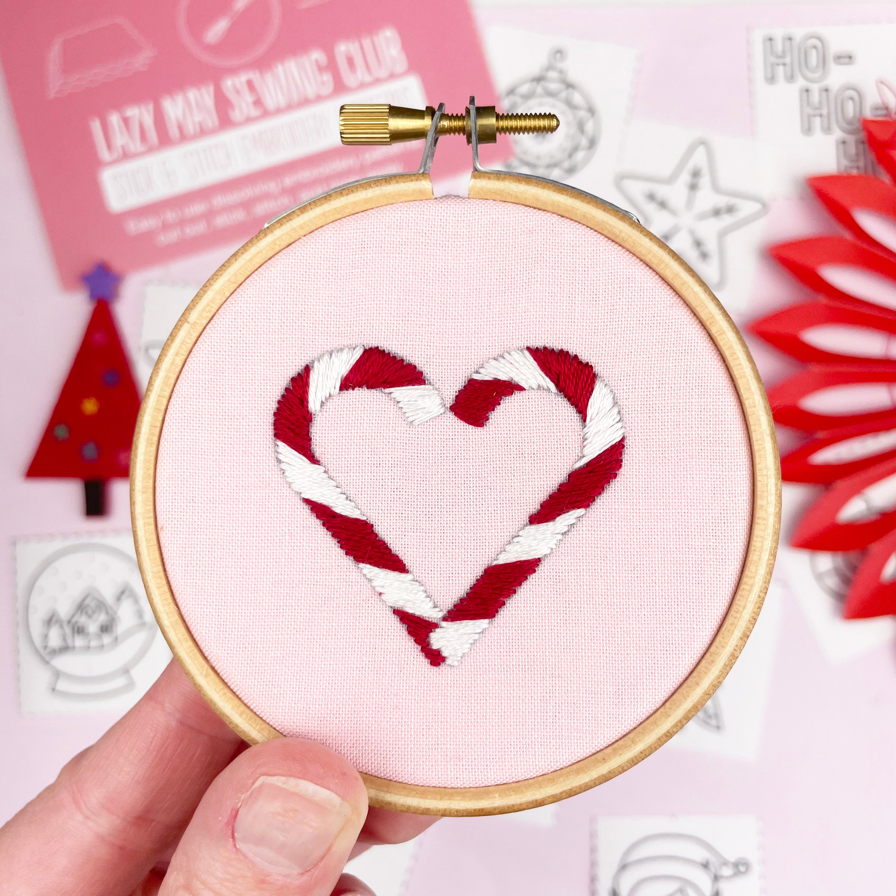 Embroidery hoop with candy cane heart design held by a hand, with a pink background and sewing club materials.