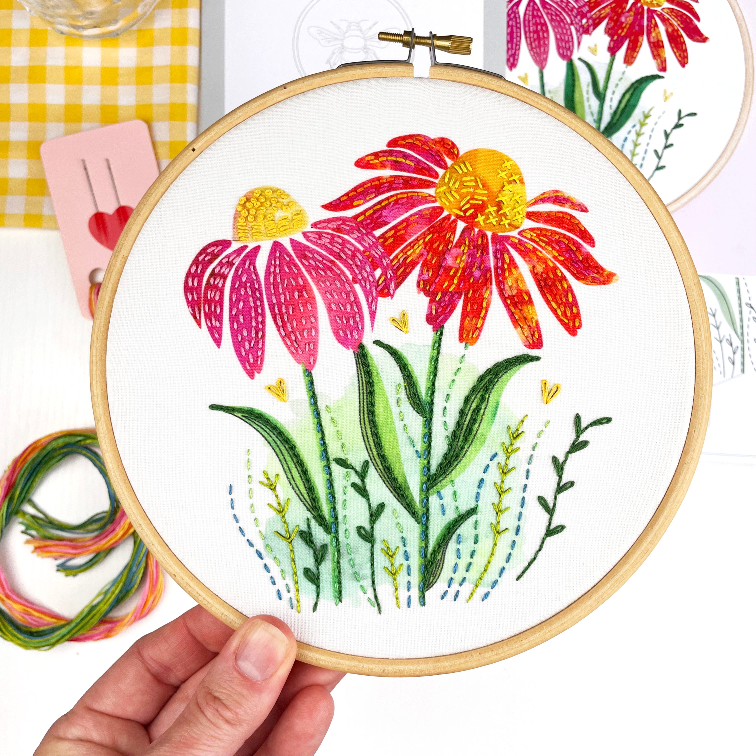 Embroidery hoop with colorful floral design held by a hand, with additional embroidery materials in the background.