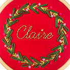 Embroidery hoop with red fabric, green and gold thread, and pink beads spelling 'Claire'.