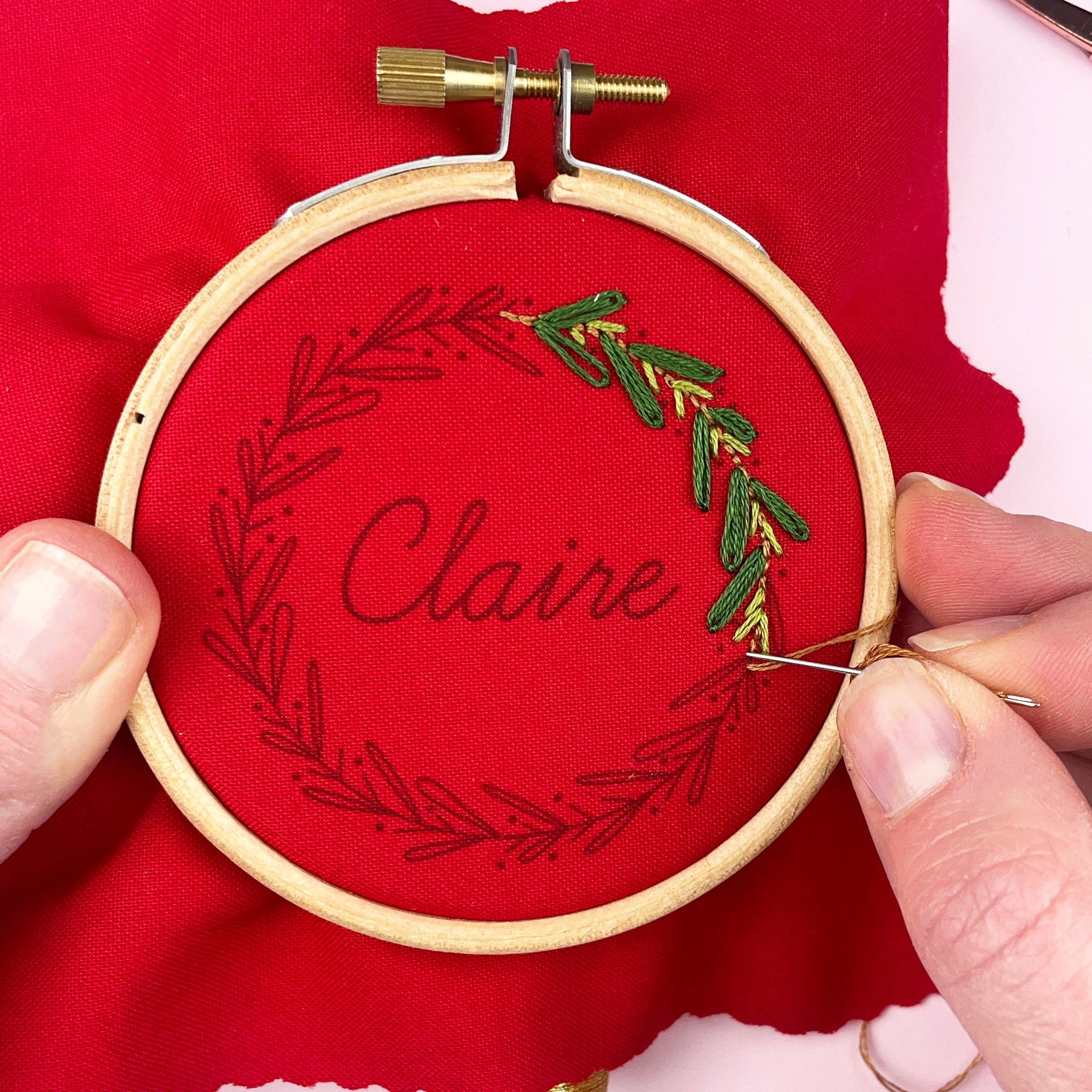 Embroidery hoop with red fabric and green leaf design, held by hands on a red background.