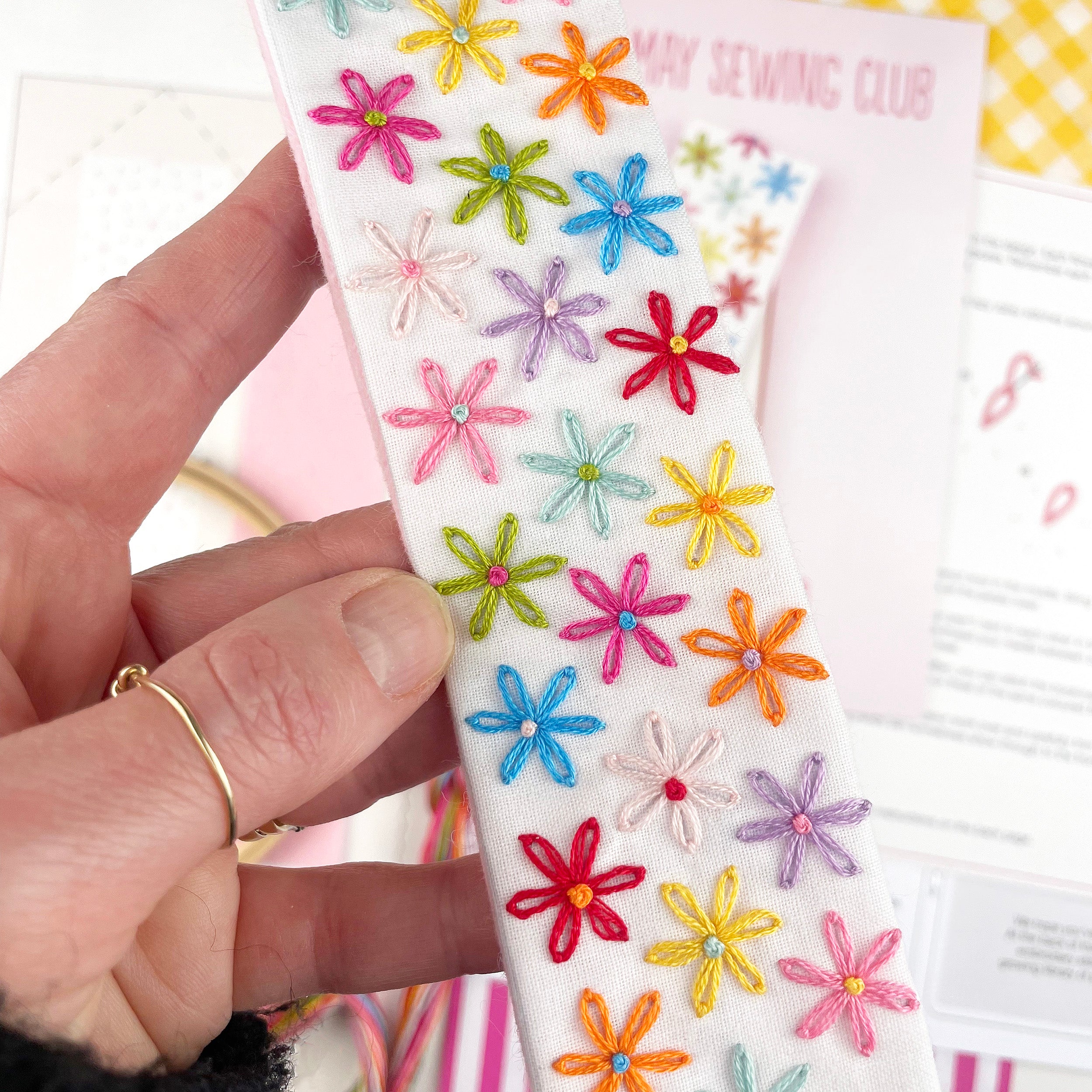 Hand holding a white fabric bookmark with colorful embroidered flowers, with 'May Sewing Club' in the background.