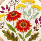 Close-up of a floral embroidery design on a white fabric background, featuring two large red flowers with yellow centers and stems, yellow flower clusters, purple buds, green leaves, and brown botanical details.
