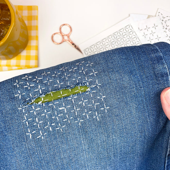 Visible mending repair on blue jeans decorated with white sashiko-inspired running stitches.