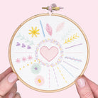 Hands holding a modern beginner embroidery hoop with colorful stitched heart and floral patterns on white fabric