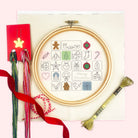 A circular embroidery hoop advent calendar kit with Christmas-themed  design on cream fabric, surrounded by crafting props including red ribbon, embroidery floss, a red card with a star, and a gold thread bundle, all arranged on a light pink background.