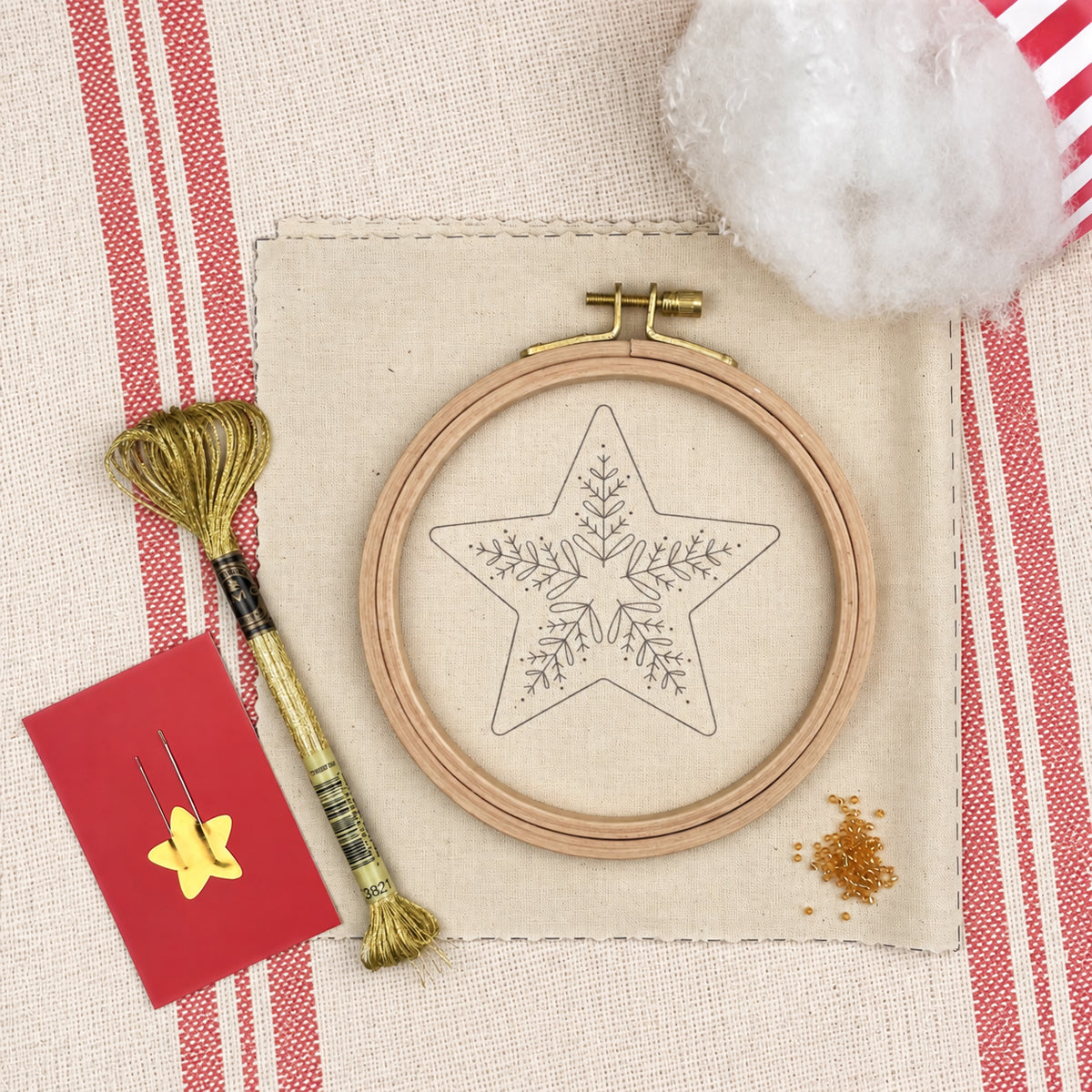 A flat-lay of embroidery supplies on a beige fabric background with red-and-white stripes. The main product appears to be a wooden hoop containing a star-shaped embroidery design, alongside a spool of gold thread, a red card with yellow star pins, loose thread, and a ball of white cotton.