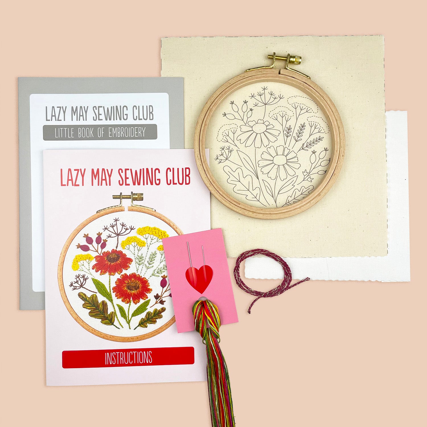 Embroidery kit components arranged on a peach background, including a wooden hoop with floral embroidery, printed instruction cards from Lazy May Sewing Club, thread, and a pink heart-shaped card.