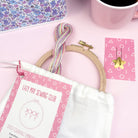 Pastel embroidery threads, wooden hoop, pins, and floral sewing bag on a soft pink surface with a pink mug nearby