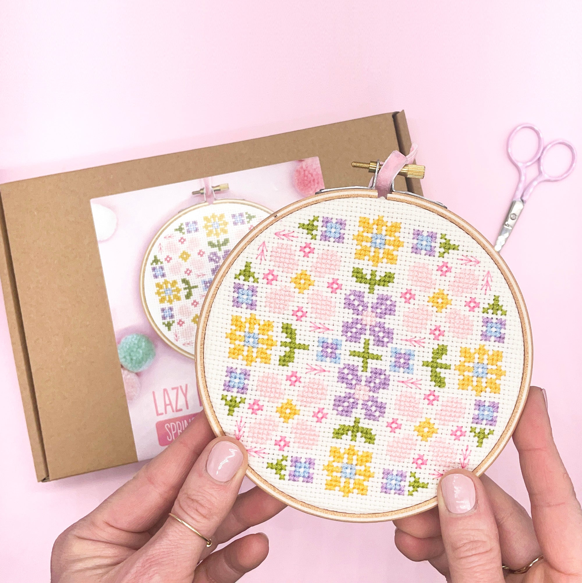 A close-up of a completed floral cross-stitch design displayed in an embroidery hoop. The pattern features a vibrant bouquet of spring flowers in shades of pink, yellow, blue, and green, showcasing intricate stitching details.