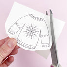 Hand holding a paper iron on embroidery transfer of a christmas jumper design with scissors on a pink background