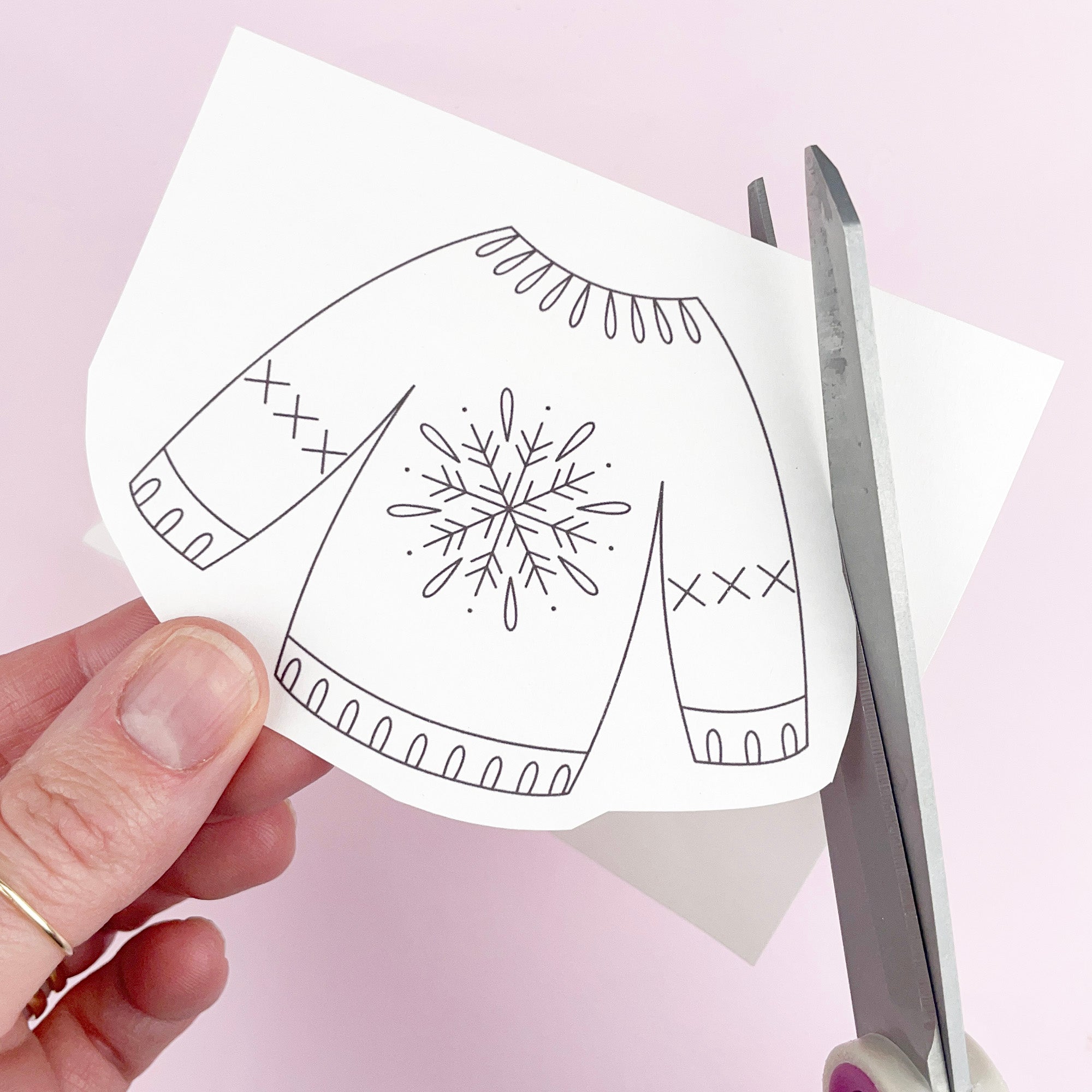 Hand holding a paper iron on embroidery transfer of a christmas jumper design with scissors on a pink background