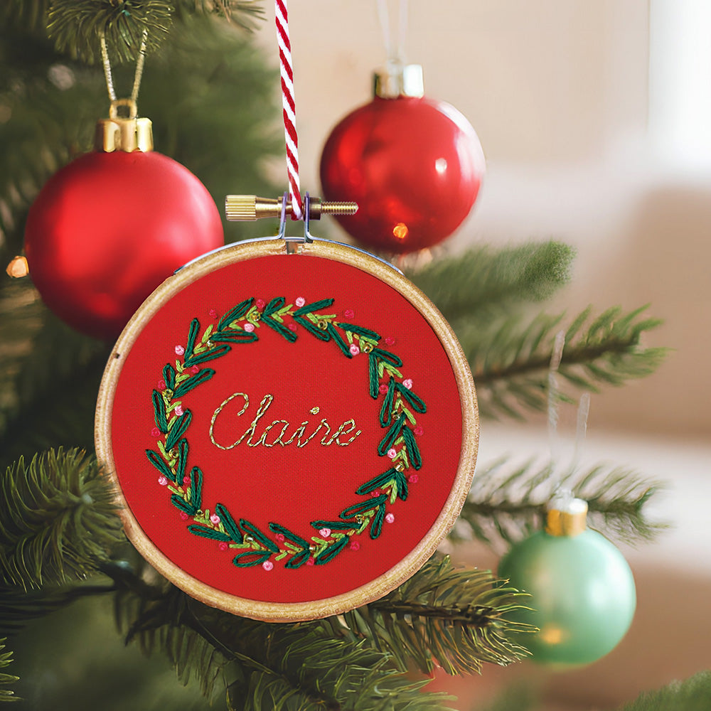 Embroidered ornament with wreath design and 'Claire' text, hanging on a Christmas tree.