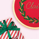 Embroidery hoop with Christmas-themed design and a wrapped gift with a green ribbon on a pink background