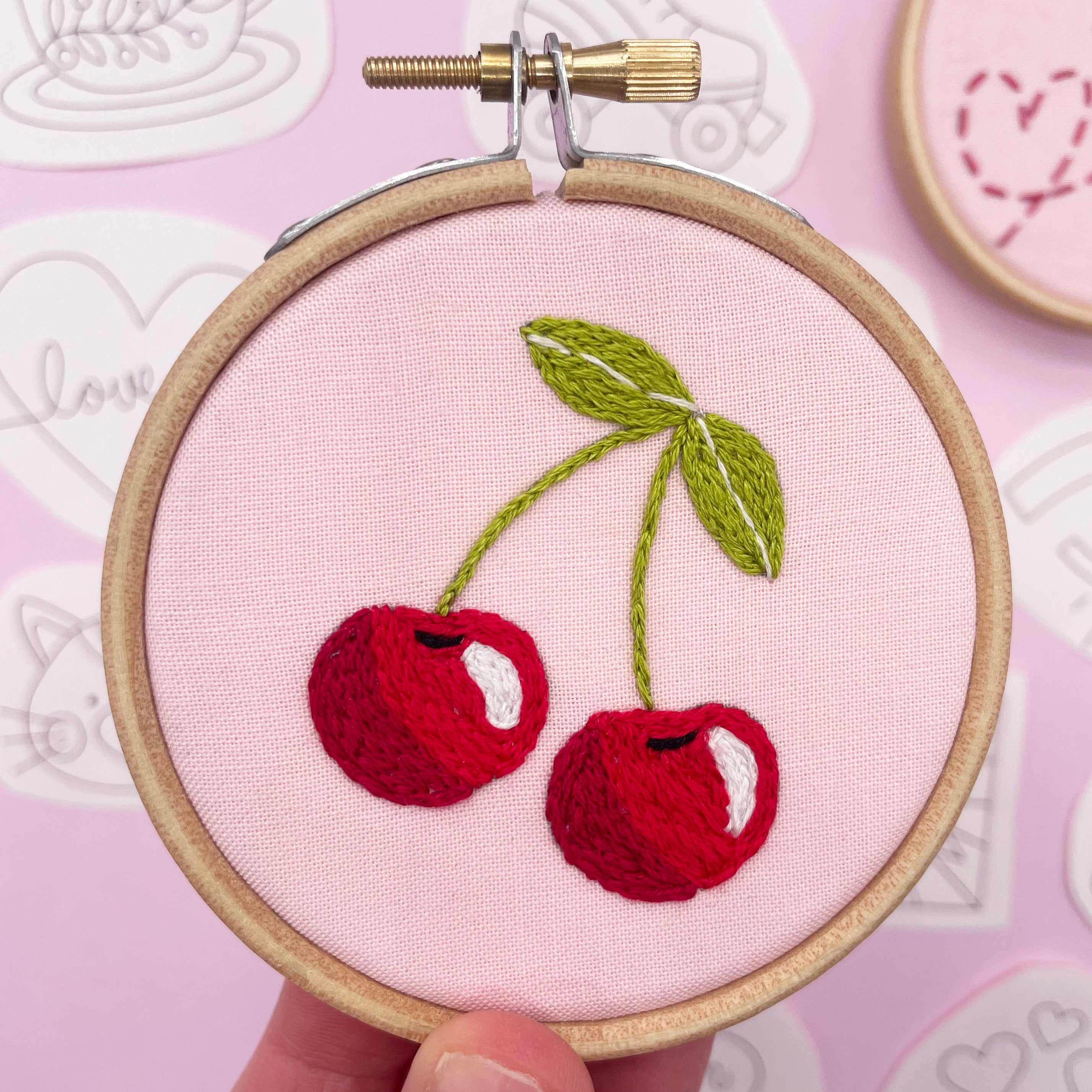 Close-up of a finished cherry embroidery design on pink fabric, stitched in a wooden hoop. A fun Easter embroidery pattern for creative stitching projects.