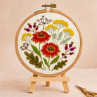 A round embroidery hoop holding a floral needlepoint or embroidered design on a small wooden stand. The design features red and yellow flowers with green leaves and purple buds on a cream fabric background.