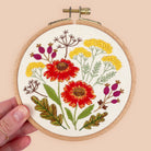 A hand holding a wooden embroidery hoop with a completed floral cross-stitch design. The embroidery features red and yellow flowers, green leaves, and purple buds on an off-white fabric background, set against a solid pale peach background.