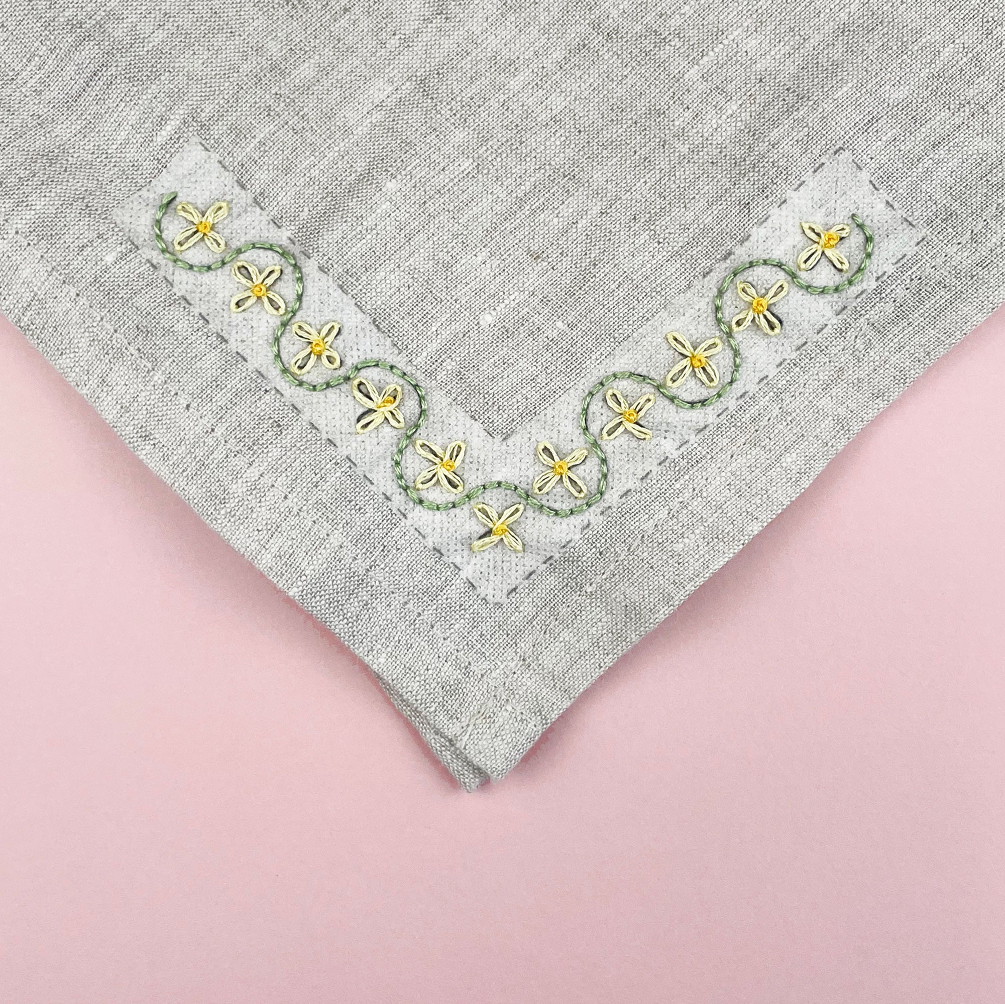 Yellow floral embroidery stitched in a corner design on the edge of a grey linen napkin.
