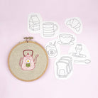 Embroidery hoop with a pink teapot design on a light background with tea-themed embroidery transfers.