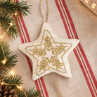 A star-shaped white fabric ornament with a gold embroidered snowflake design, hanging on a red and beige striped textile with pine branches and warm string lights around it.