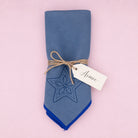 A folded dusty blue fabric napkin with a dark blue embroidered star design and a solid royal blue pointed tip, tied with twine and a white paper tag that reads 'Aimée', set against a solid pink background.
