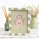 Framed embroidered picture of a pink teapot with a flower on a checkered tablecloth.