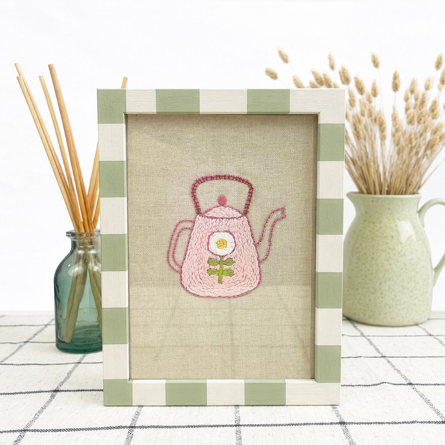 Framed embroidered picture of a pink teapot with a flower on a checkered tablecloth.