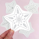 A hand holds a white star-shaped embroidery pattern with black line-art snowflake and floral designs, with several similar star patterns blurred in the background on a soft pink surface.