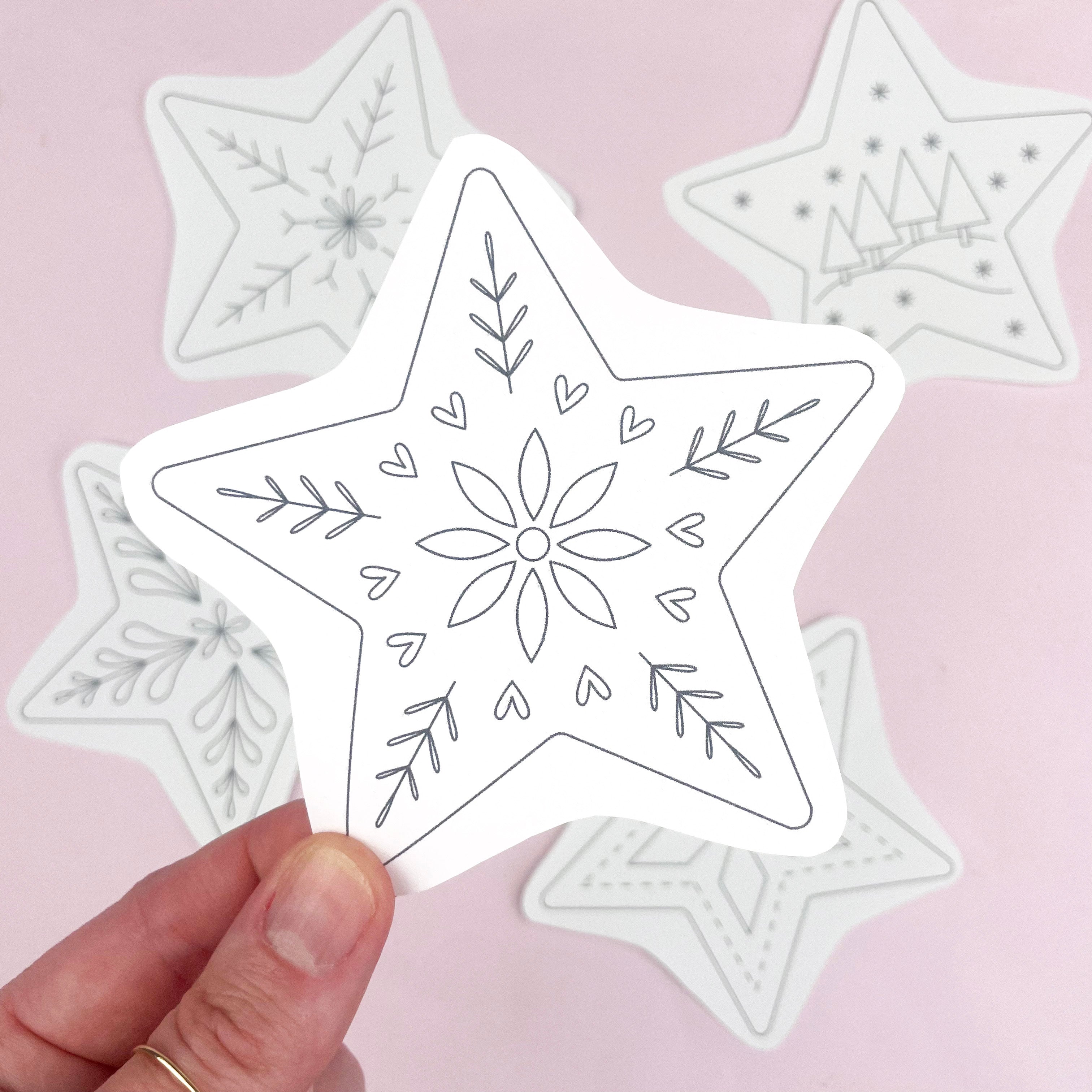 A hand holds a white star-shaped embroidery pattern with black line-art snowflake and floral designs, with several similar star patterns blurred in the background on a soft pink surface.