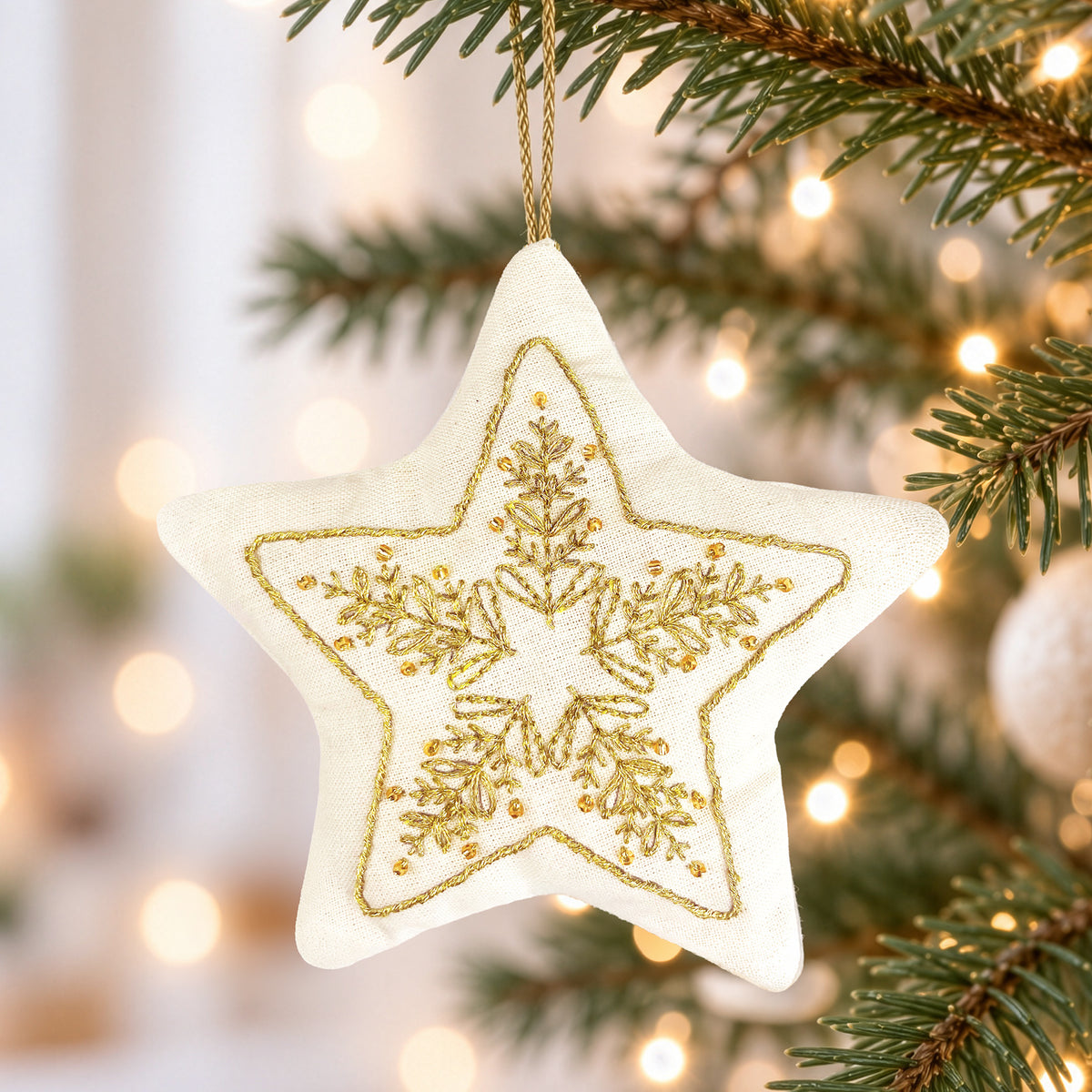 A cream-colored fabric star ornament with a gold embroidered snowflake design, hanging from a gold string on a Christmas tree branch with warm bokeh lights in the background.