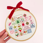 A hand holds a wooden embroidery hoop advent calendar containing a completed Christmas-themed embroidery design with embroidered icons, a red ribbon bow, and a 'Merry Christmas' message on a light pink background.