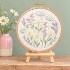 A round embroidery hoop holding a floral embroidered design on cream fabric, displayed on a small wooden stand. The embroidery features white daisies, pink and purple flowers, and green stems on a light wood background.