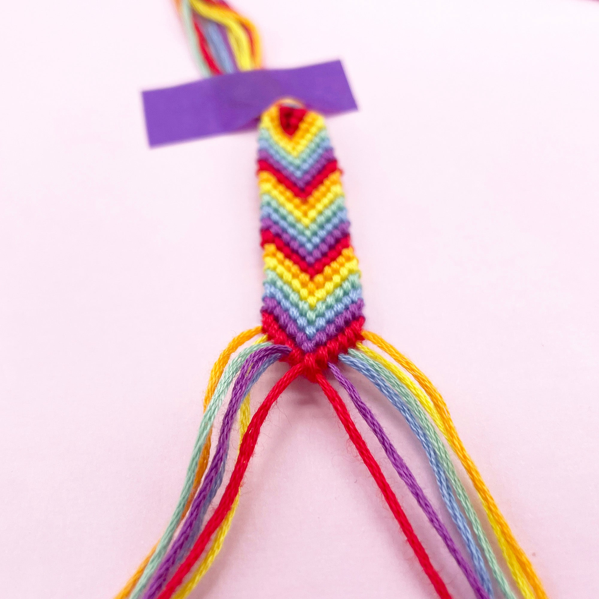 A rainbow friendship bracelet in progress, with colourful embroidery thread strands taped down on a pastel background. DIY bracelet making kit for beginners.