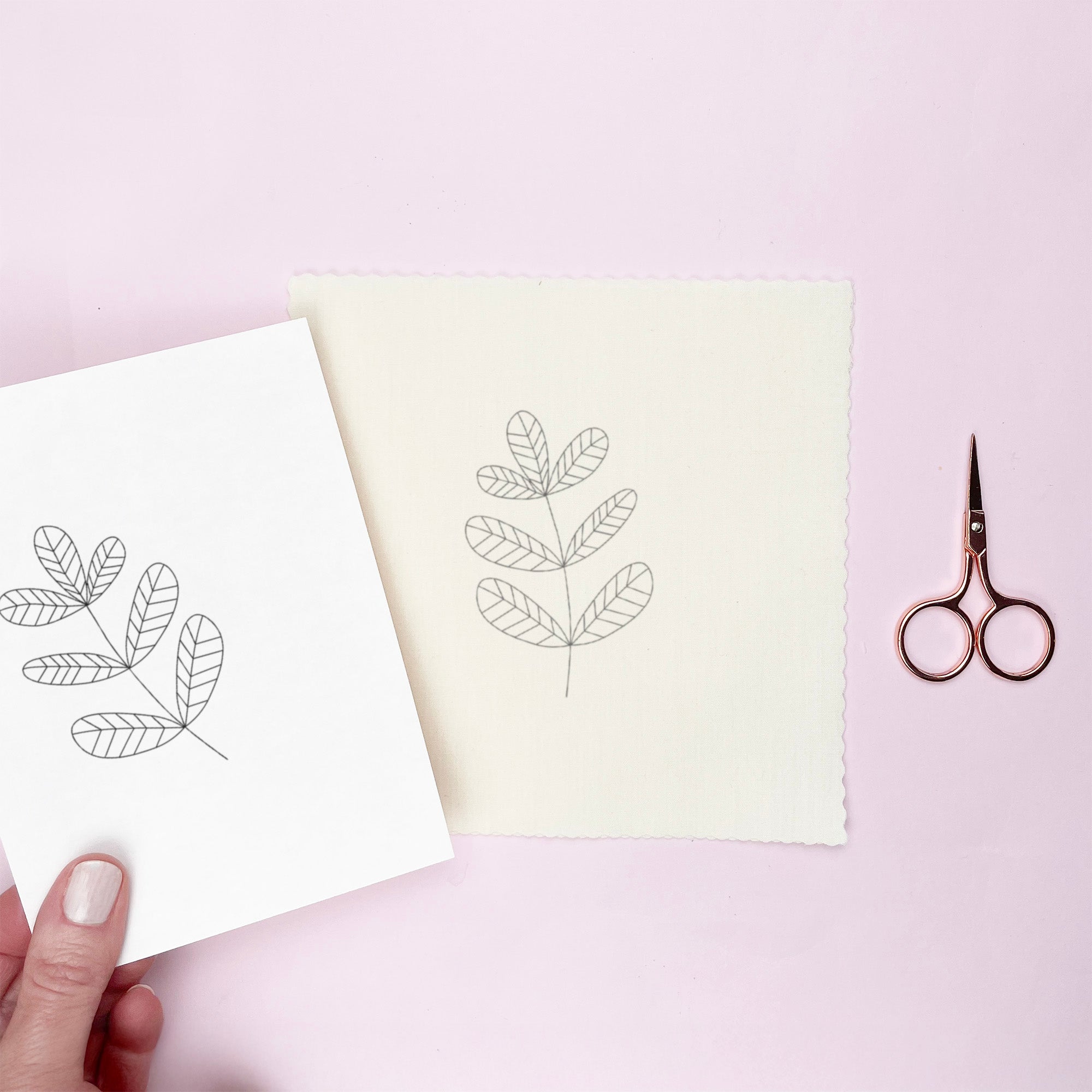 Hand holding a piece of transfer paper with leaf designs on a light pink background with scissors.