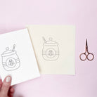 Hand iron on embroidery transfer with a honey jar design on a light pink background with scissors.