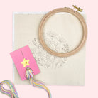 A flat-lay of embroidery supplies on a pale pink background, including a wooden embroidery hoop, a beige fabric sheet with a floral line drawing, a pink card holding a yellow star-shaped pin, and strands of multicolored embroidery thread.