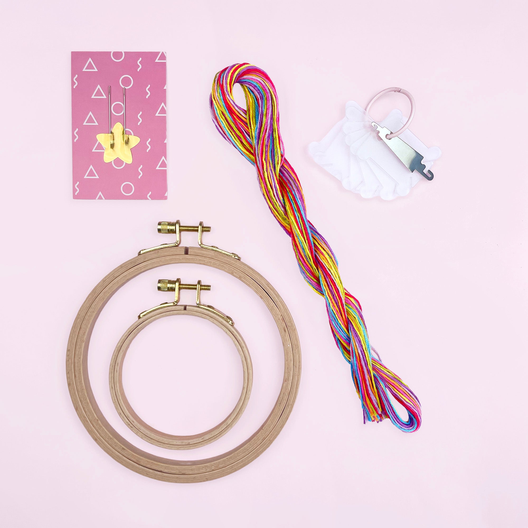 Two wooden embroidery hoops, colorful threads, needle set, and small tools on a pink background