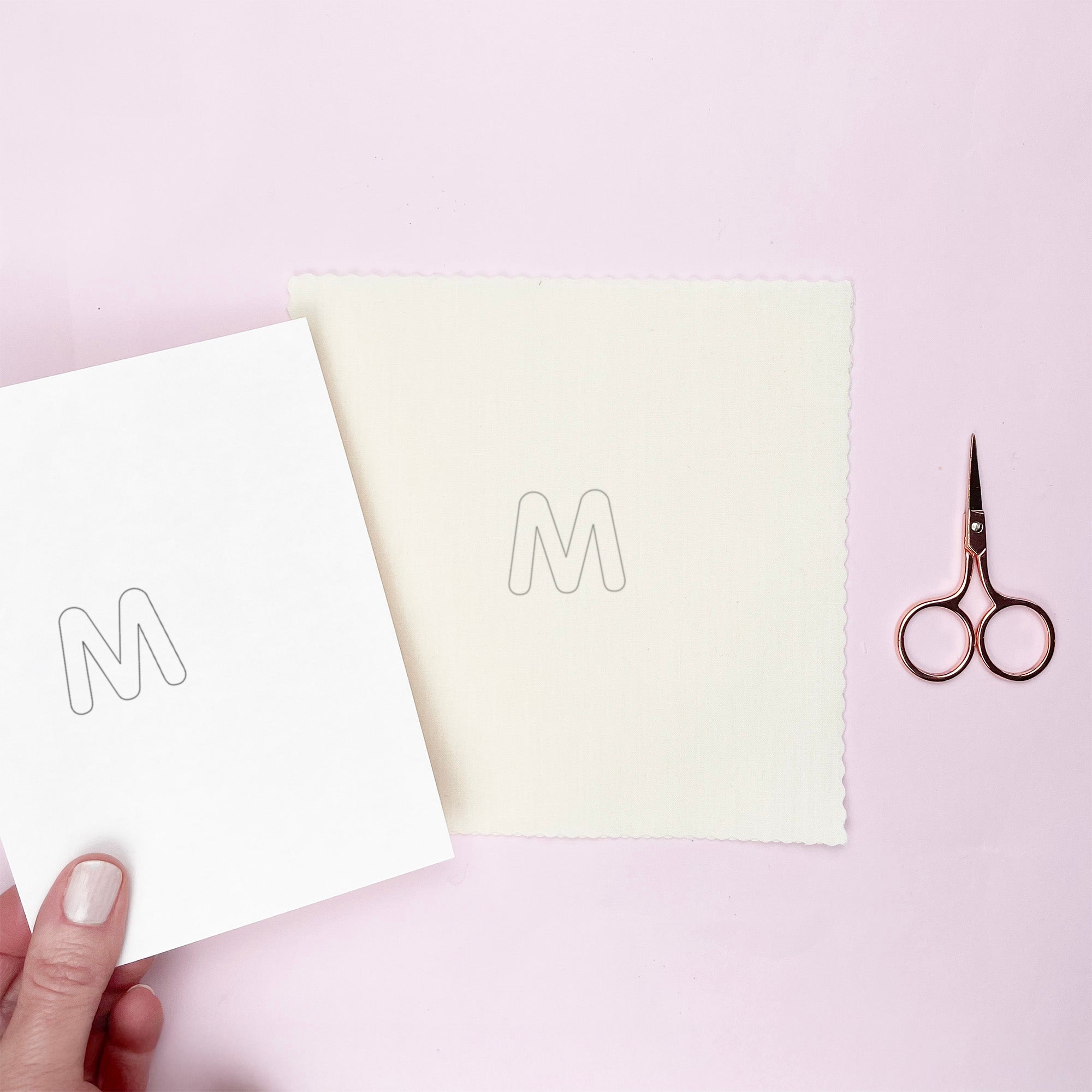 Hand holding a piece of paper with letter 'M' cutout, next to a iron on embroidery pattern with a similar cutout and a pair of scissors on a light pink background.