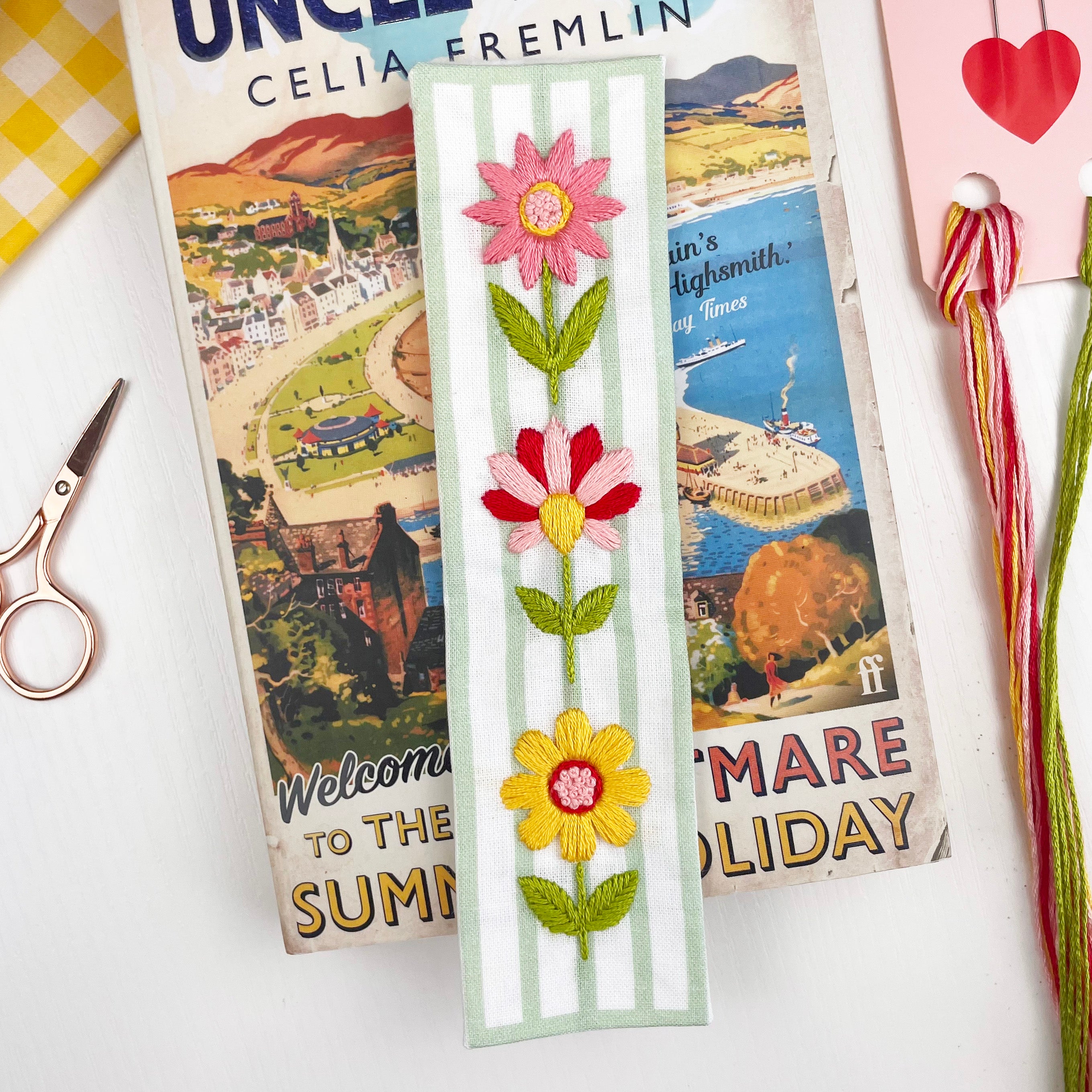 Floral bookmark with vintage book covers in the background. In the image you can see embroidery thread and scissors, part of a make your own bookmark kit