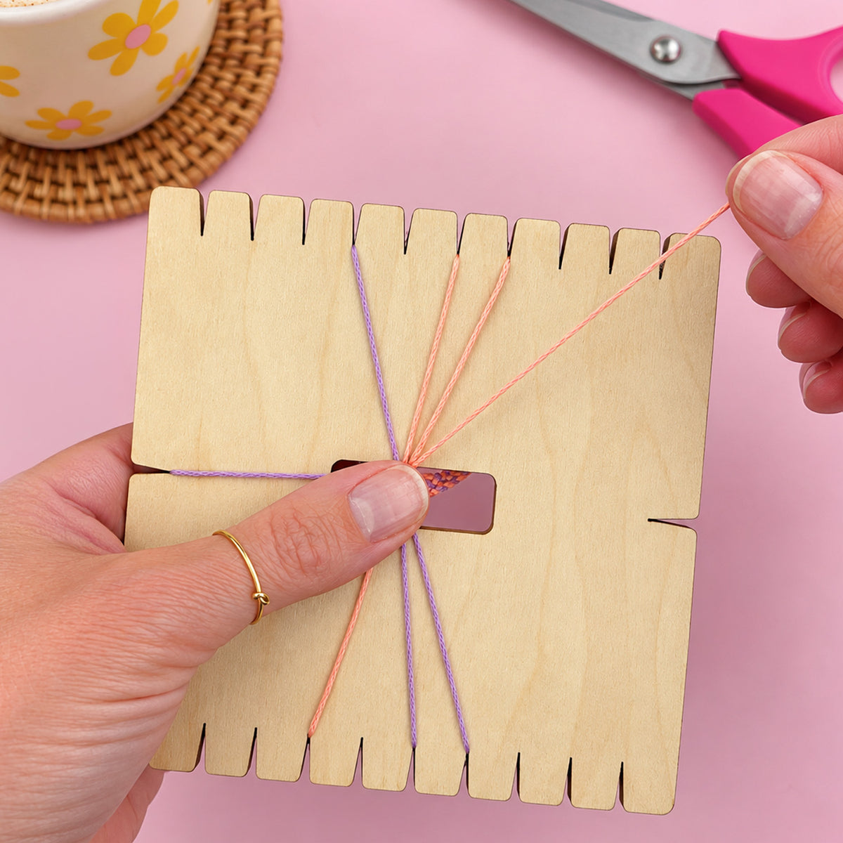 A close-up of hands using a wooden loom needle to weave peach and purple yarn, with pink scissors visible in the background on a pink surface.