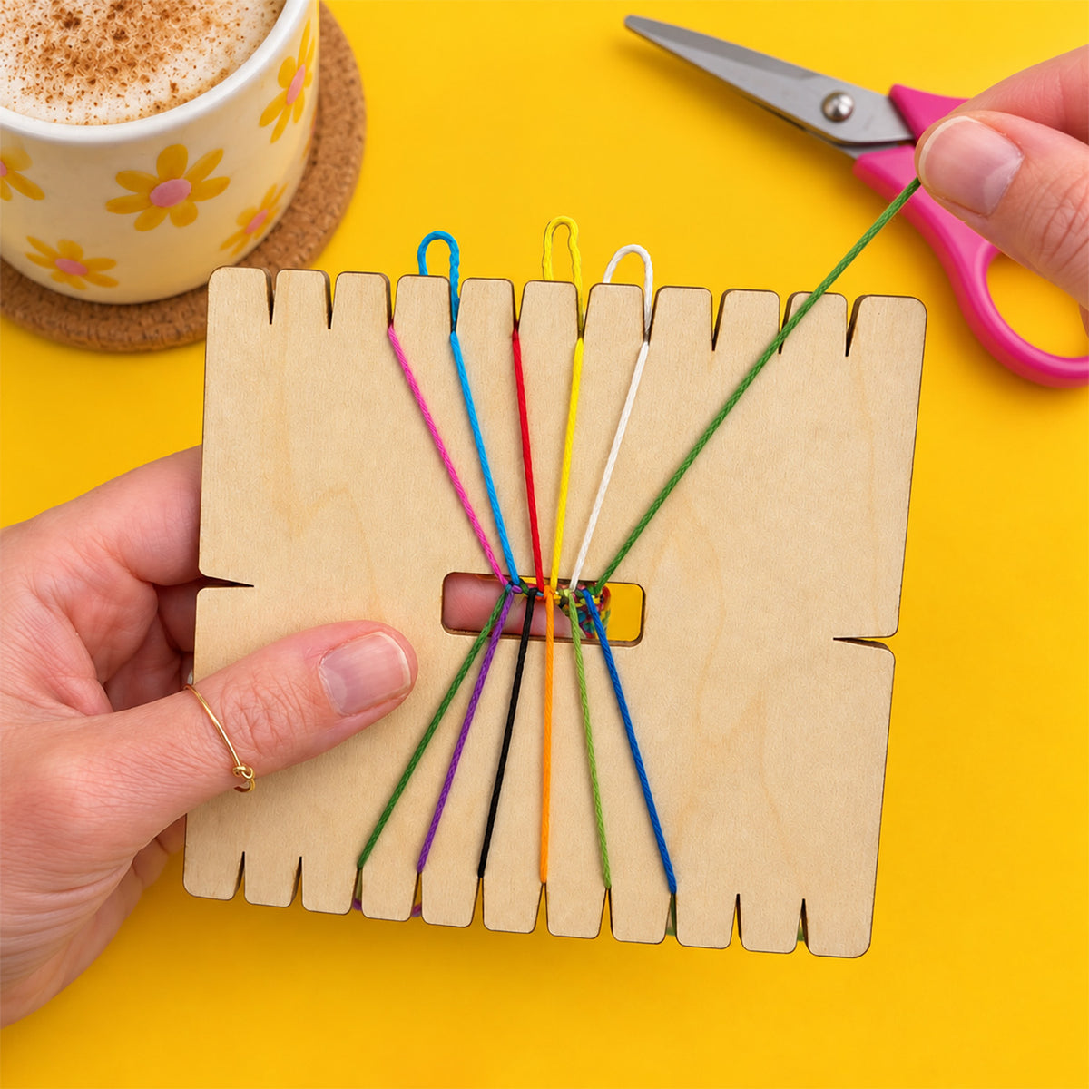 A wooden loom card being held by hands, with colorful thread needles threaded through its slots. A hand holds a green thread needle, and pink-handled scissors and a coffee cup are visible in the background on a bright yellow surface.