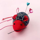 Red fabric mouse with a blue patterned body, black eyes, and pink hair on a light pink background. cotton mouse pin cushion with embroidery pins, ideal for sewing, quilting, and craft projects.

